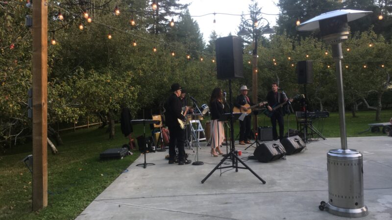 Band playing outside the hall at Mitchell Grove near dusk, surrounded by orchard and redwoods.